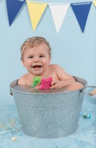 One year old boy in a metal bath tub playing with a toy and smiling at the camera